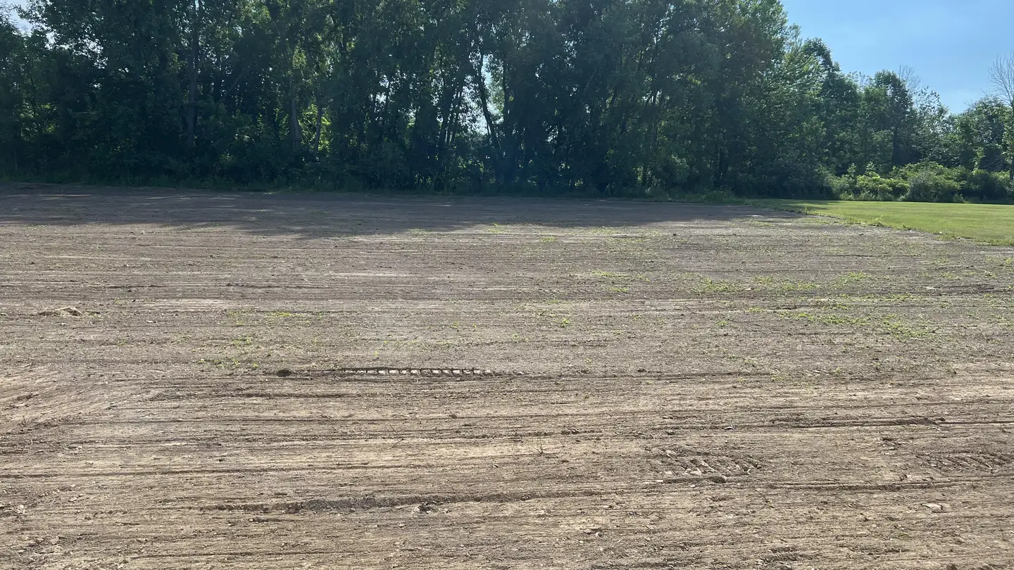 Wide, flat lot graded smooth with machine tracks in the dirt and a hardwood treeline behind