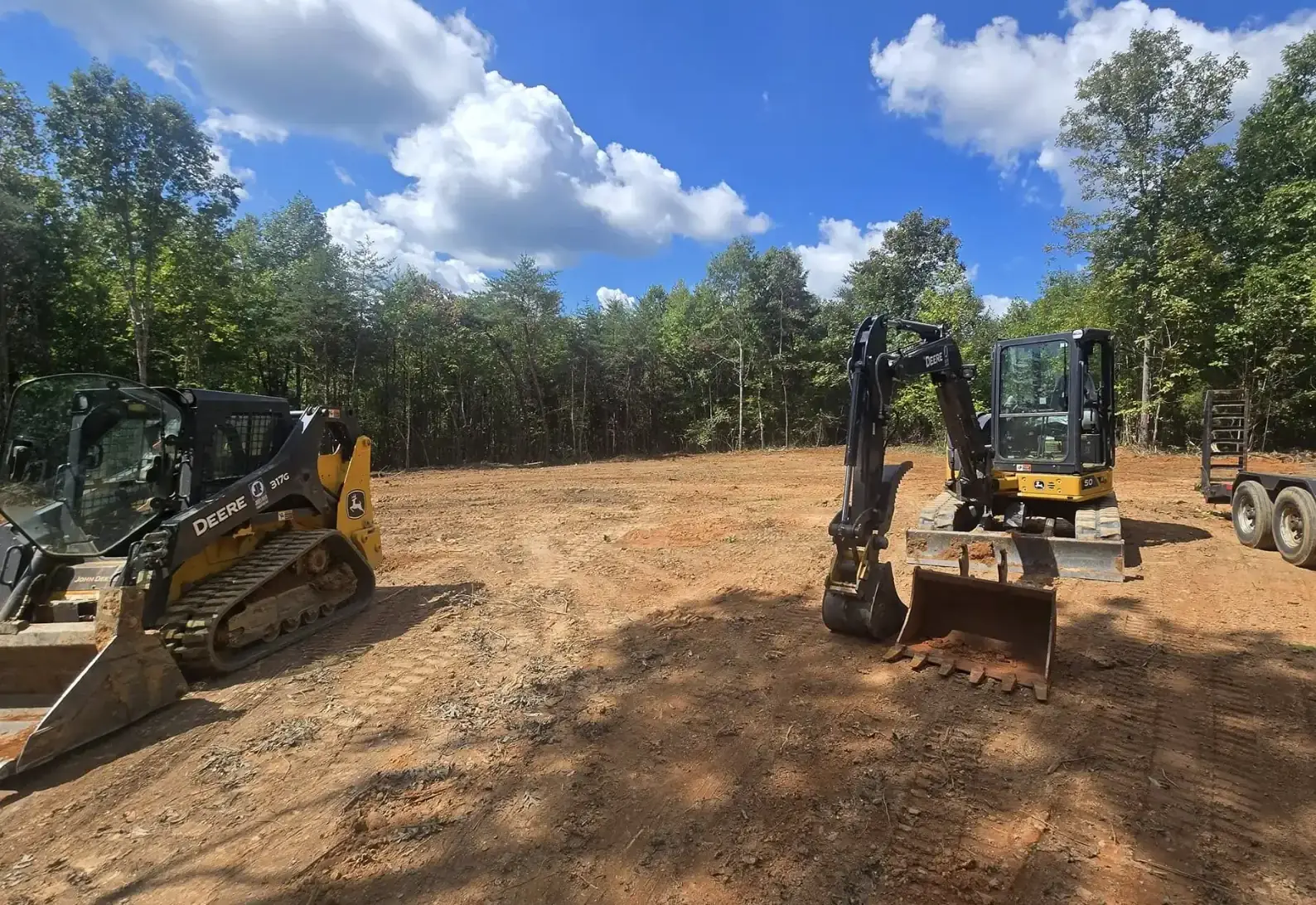 Compact track loader and mini excavator parked on a freshly cleared lot ringed by woods