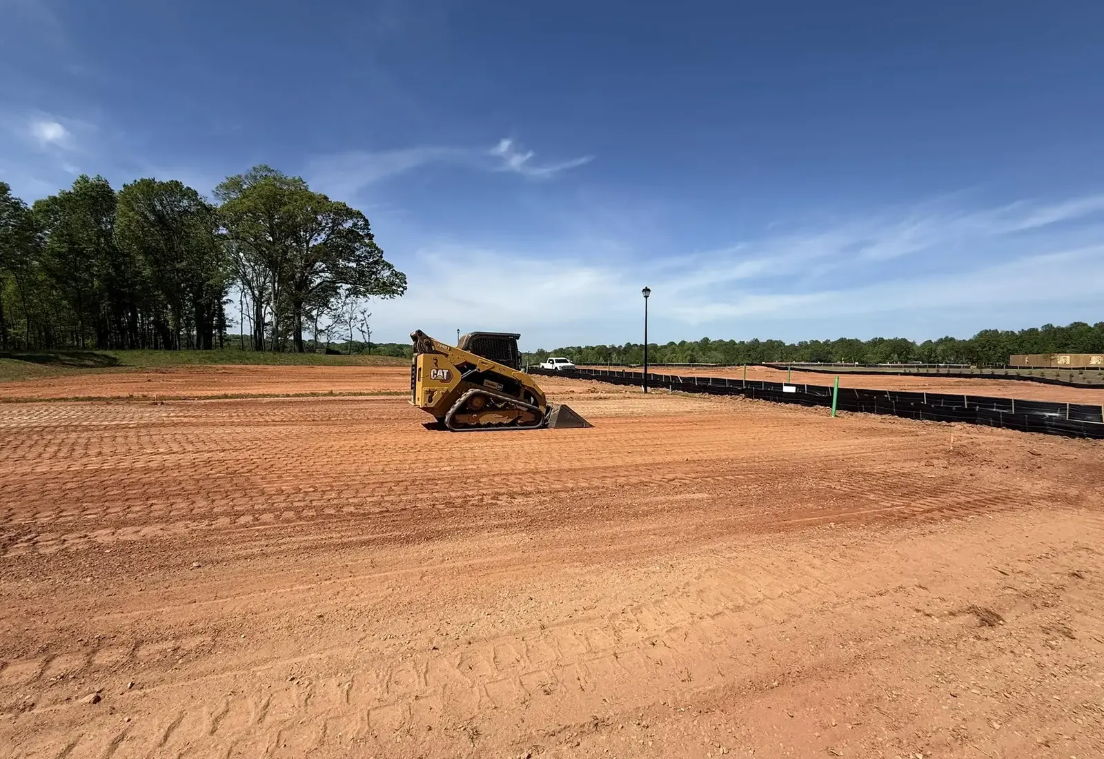 Compact track loader on a wide red clay pad graded smooth with silt fence along the treeline
