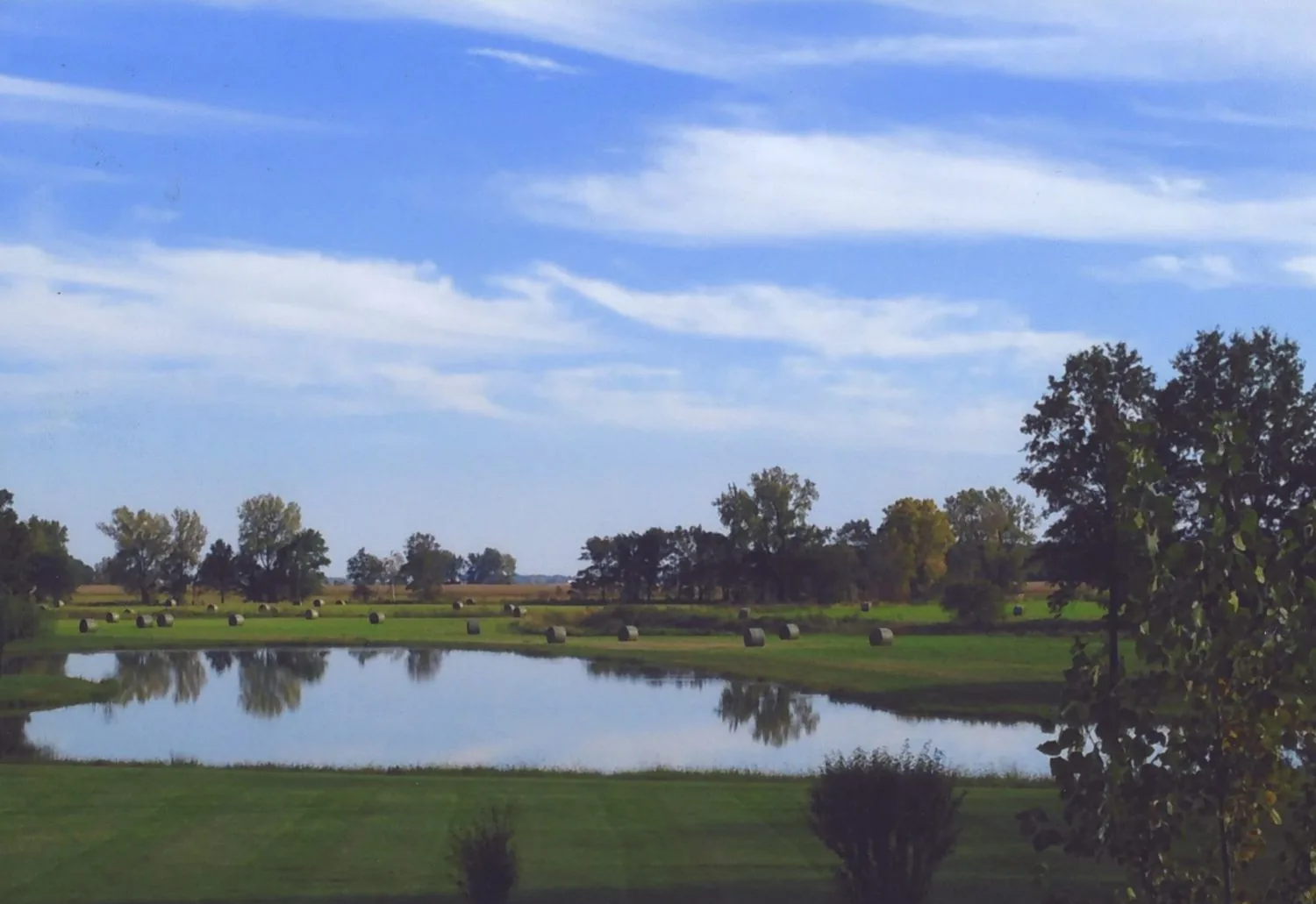 Full farm pond on open pasture with round hay bales and a hardwood treeline beyond